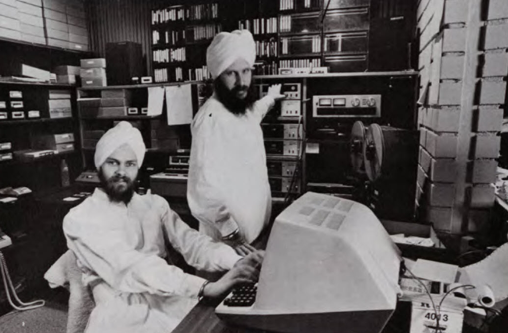 Two 3HO
													 members in all white circa 1986 hanging out in a media studio, one
													 sitting at a computer and the other standing in front of some recording
													 equipment.